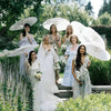 Un grupo de mujeres con vestidos elegantes sostienen parasoles de papel blancos en un jardín al aire libre.