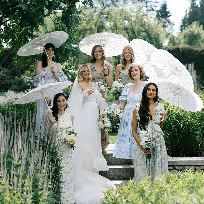 Un grupo de mujeres con vestidos elegantes sostienen parasoles de papel blancos en un jardín al aire libre.