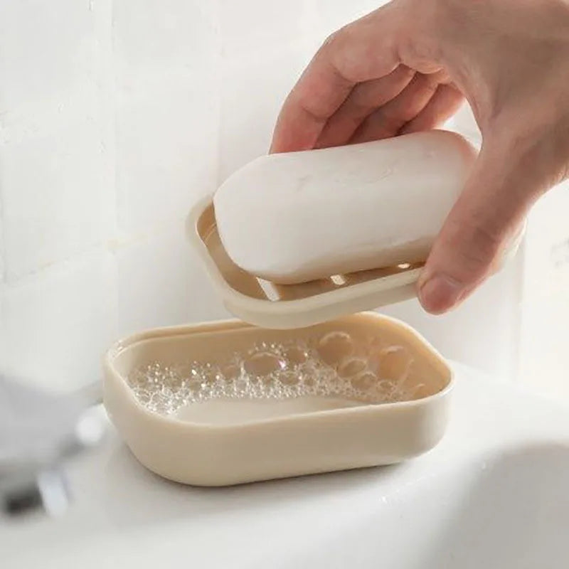 A hand is holding a white double layer drain soap dish, with a filled beige soap dish nearby.