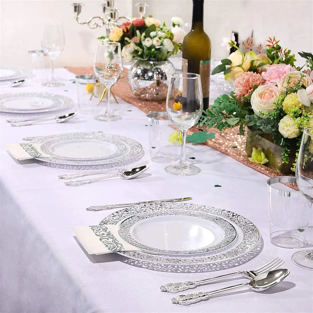 Silberbesteck mit Spitze für Partygeschirr auf weißem Tischtuch mit Blumenarrangements.