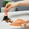 A hand is holding an orange mini broom and dustpan set on a white desk surface.