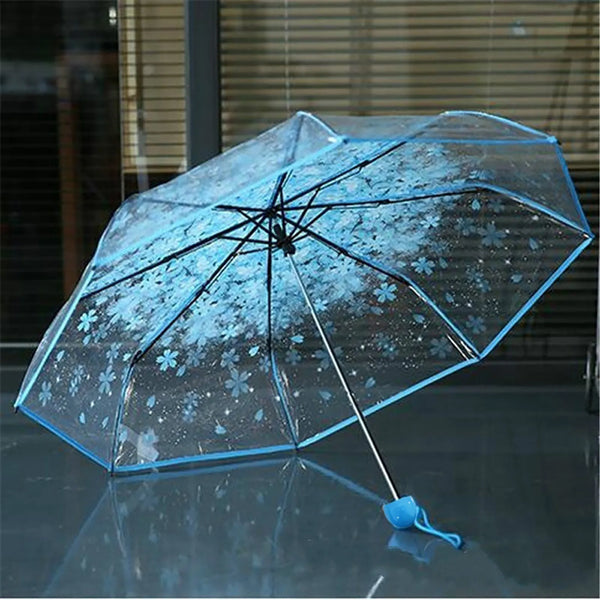 A clear folding umbrella with a cherry blossom print is shown open on a reflective surface.