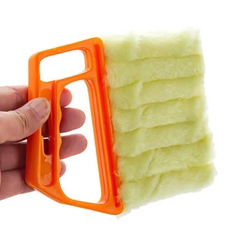 An orange multi-functional cleaning brush with a yellow fuzzy head is held by a hand.