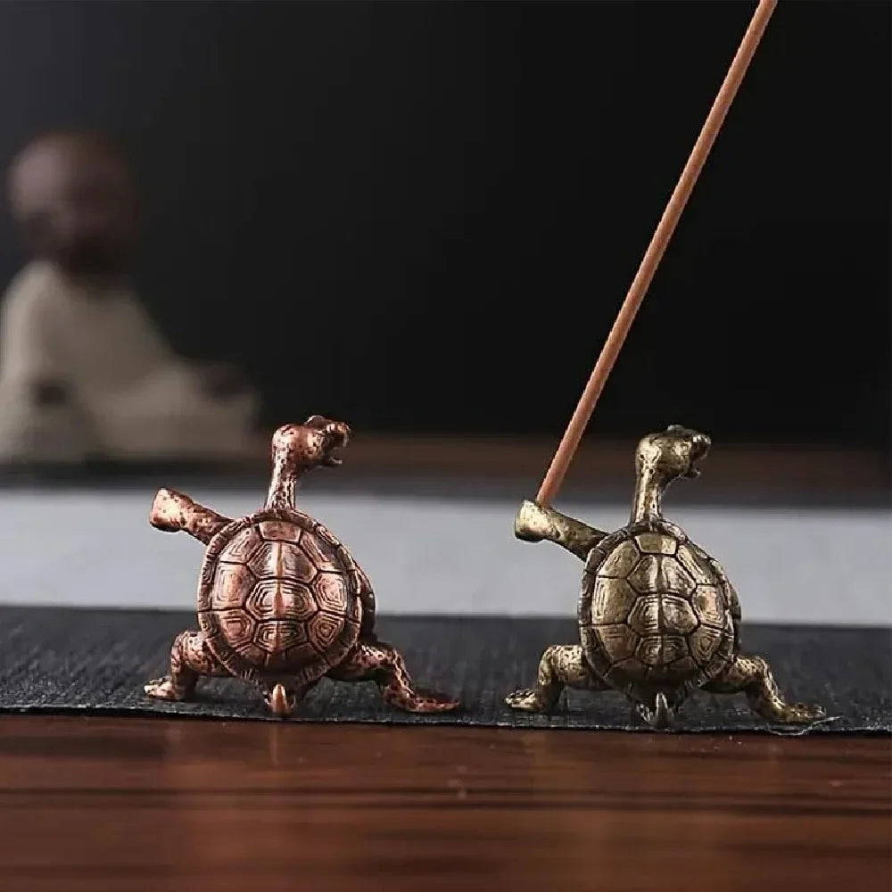 Une petite figurine de tortue en métal antique est à côté d'un bâton en bois mince sur une surface sombre.