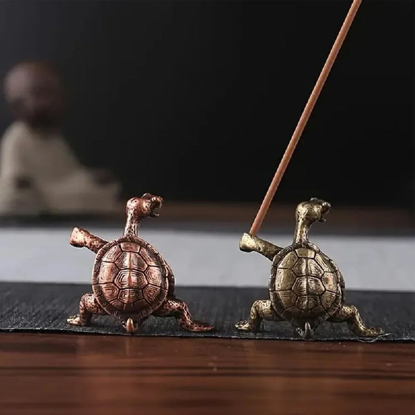 A small, antique-style metal turtle figurine is shown next to a thin wooden stick on a dark surface.