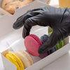 A hand in a black nitrile glove is placing pink, blue, and yellow macarons into a white box.