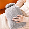 A hand is holding a large, fluffy gray plush hot water bottle against a beige knitted fabric.
