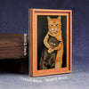 Gerahmtes Gemälde mit einer orange-weißen Katze, die eine schwarze Katze hält, in einem Holzrahmen.