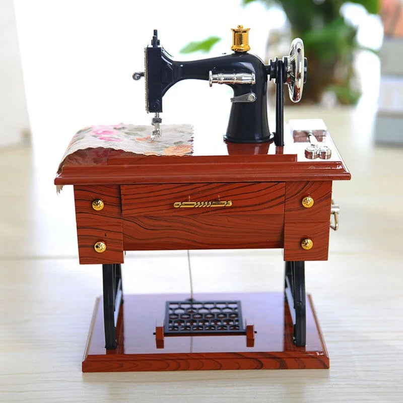 A vintage clockwork sewing machine music box with a dark body, gold accents, and a wooden base.
