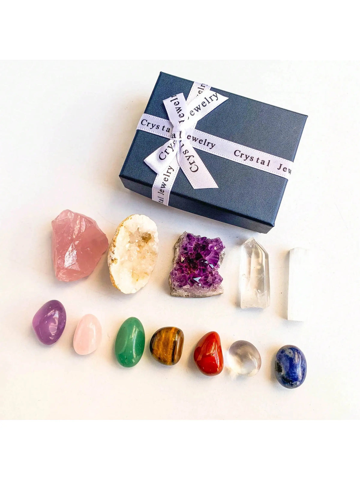 A dark blue box with white ribbon packaging holds a set of natural crystal stones including pink, yellow, purple, green, ambe