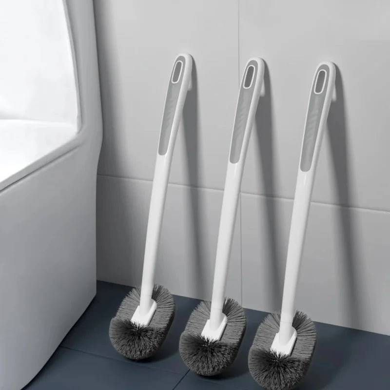 A white wall-mounted toilet brush with an ergonomic long handle is shown against a gray tiled wall.