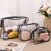 A set of three clear plastic travel makeup organizer bags resting on a light surface next to a vase of white flowers.