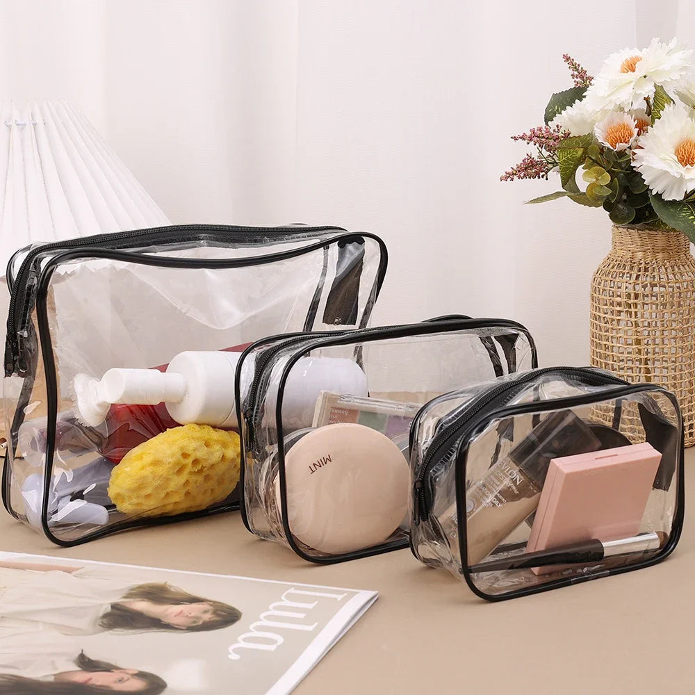 A set of three clear plastic travel makeup organizer bags resting on a light surface next to a vase of white flowers.