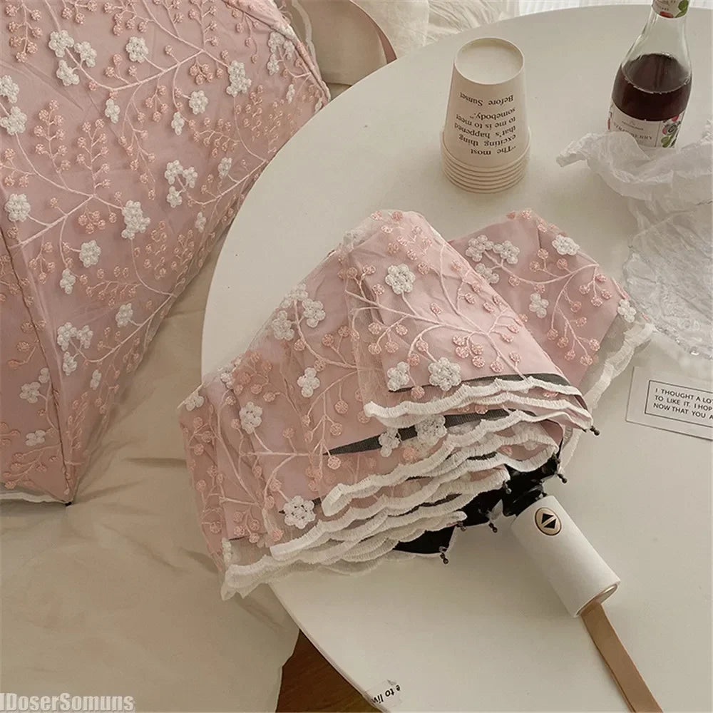 A pink embroidered sun umbrella is displayed on a white table next to a glass bottle and white fabric.