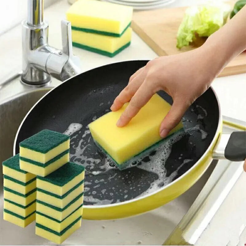 A hand is using yellow and green double-sided scouring sponges in a black frying pan for kitchen cleaning.