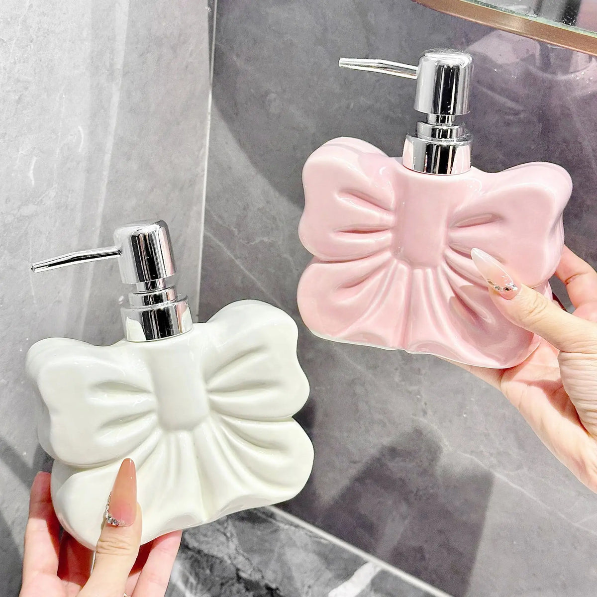 A hand holds a pink ceramic pump dispenser bottle with a bowknot design next to a white one on a marble surface.