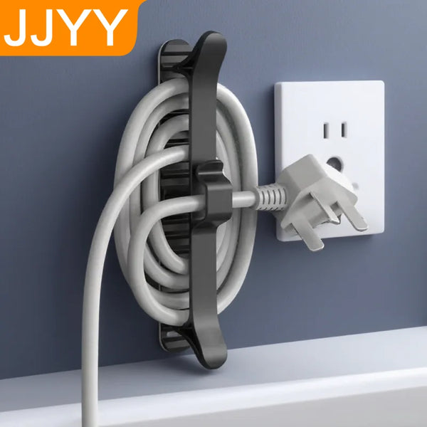 A black cable organizer with an adhesive mount is shown mounted on a gray wall next to a white electrical outlet.