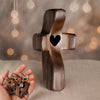 A handcrafted wooden cross with a heart cutout is displayed on a surface, next to a pile of small wooden crosses.