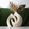 A modern minimalist ceramic vase holds dried grasses and a fan leaf arrangement against a dark green backdrop.