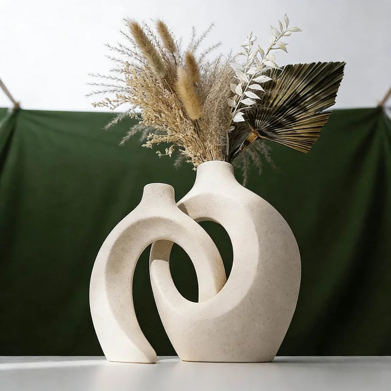 A modern minimalist ceramic vase holds dried grasses and a fan leaf arrangement against a dark green backdrop.