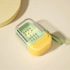 A leakproof travel container in light green and yellow plastic with a silicone strap, resting on a beige surface.