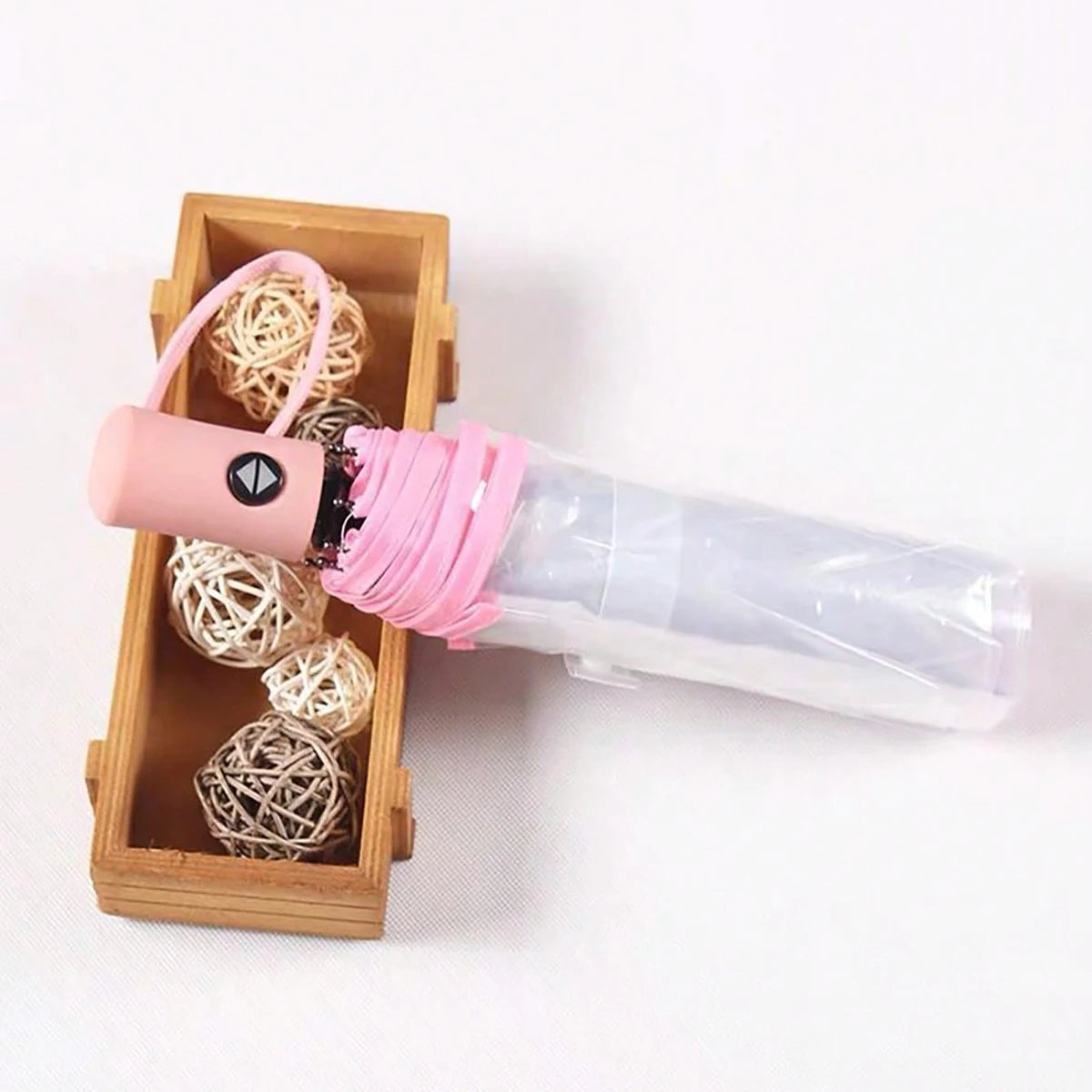 A pink automatic transparent folding umbrella with a cork handle and coiled cord is shown in a wooden box.