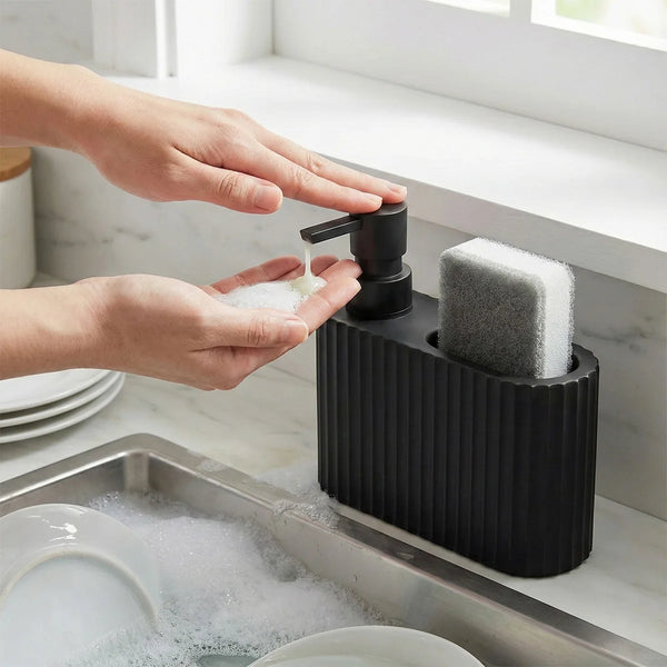 Hands dispense dish soap from a black dispenser into a stainless steel sink filled with soapy water and plates.