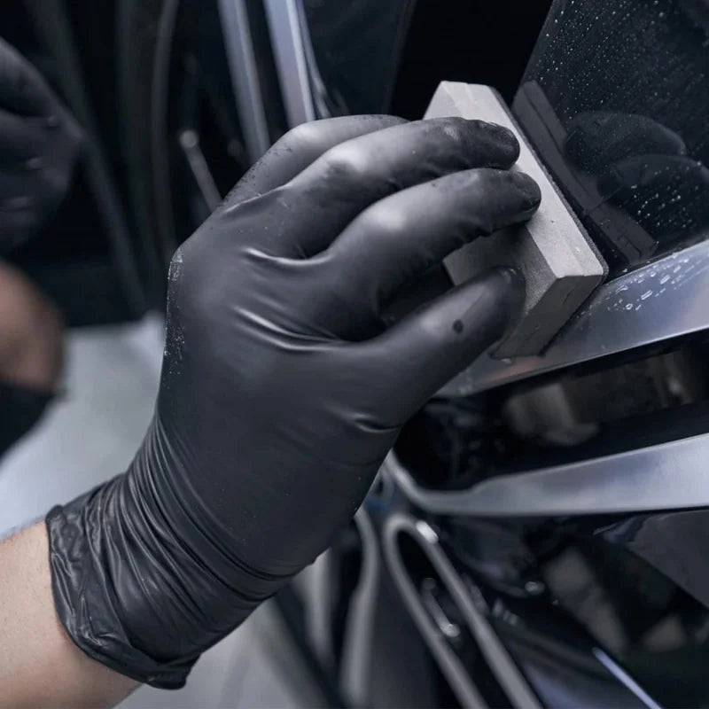 A hand wearing a black disposable nitrile cleaning glove is placing a wooden block onto a dark surface.