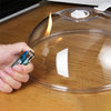 A hand places a white paper towel onto a transparent splash-proof oven cover while a flame is visible beneath it.