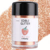 A glass jar of edible glitter powder in rose gold color with a strawberry illustration against a background of orange glitter
