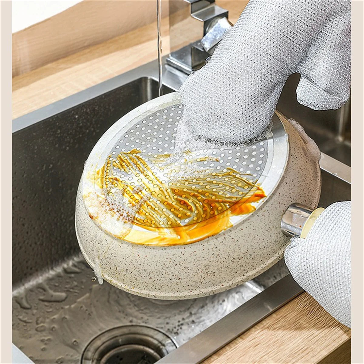 A hand wearing steel wire dishwashing gloves is washing dishes in a stainless steel sink.