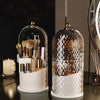 A clear glass organizer for makeup brushes and tools with a rotating lid, featuring a white base.