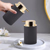 A hand is holding a black foaming soap dispenser with a gold cap, set on a white marble surface.