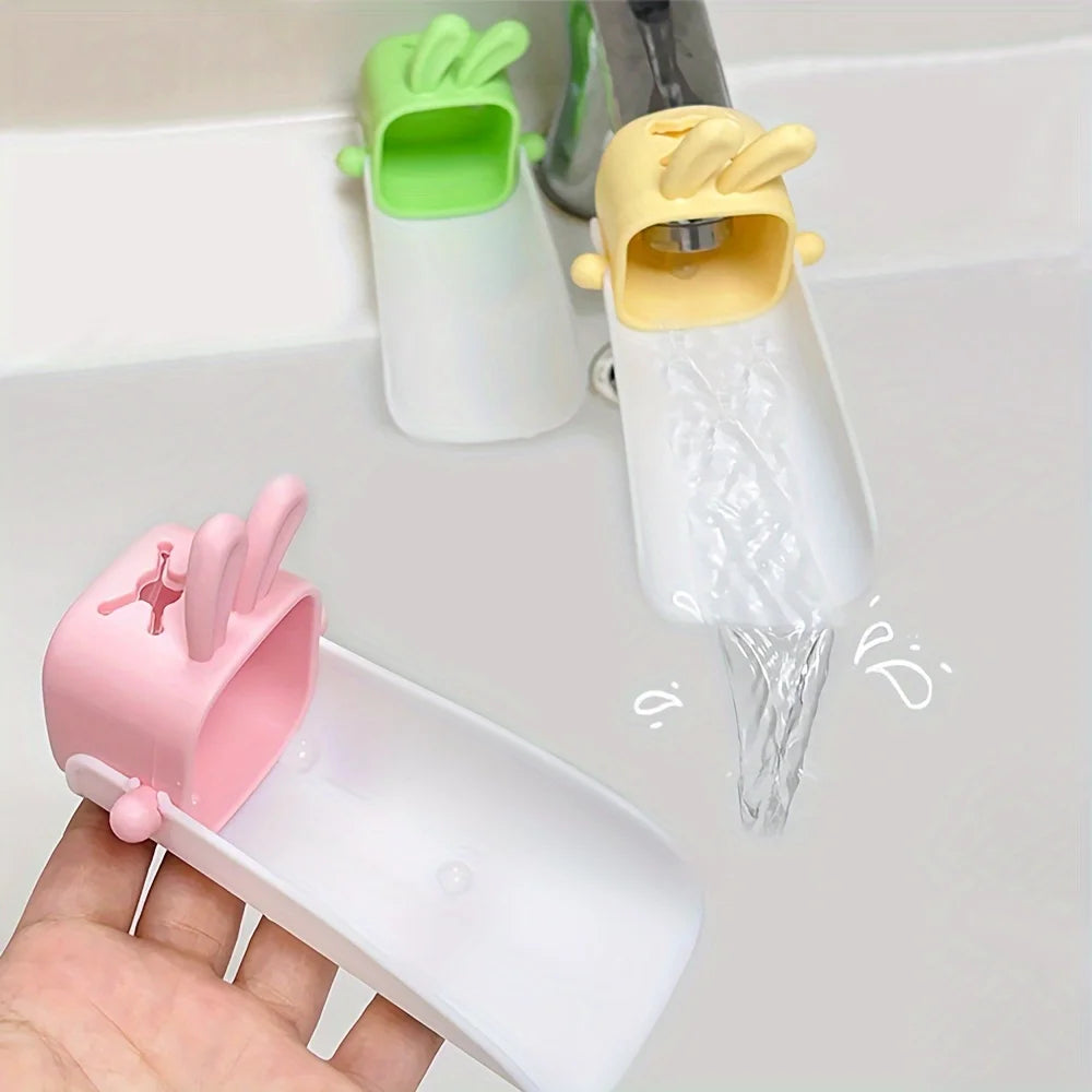 A hand holds a white faucet extender with a pink rabbit design, next to two green and yellow faucet extenders.