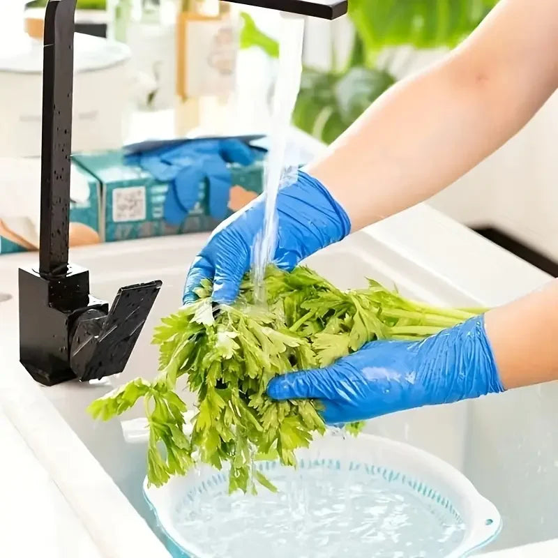A hand wearing blue nitrile disposable gloves is washing fresh green herbs under a faucet in a kitchen sink.