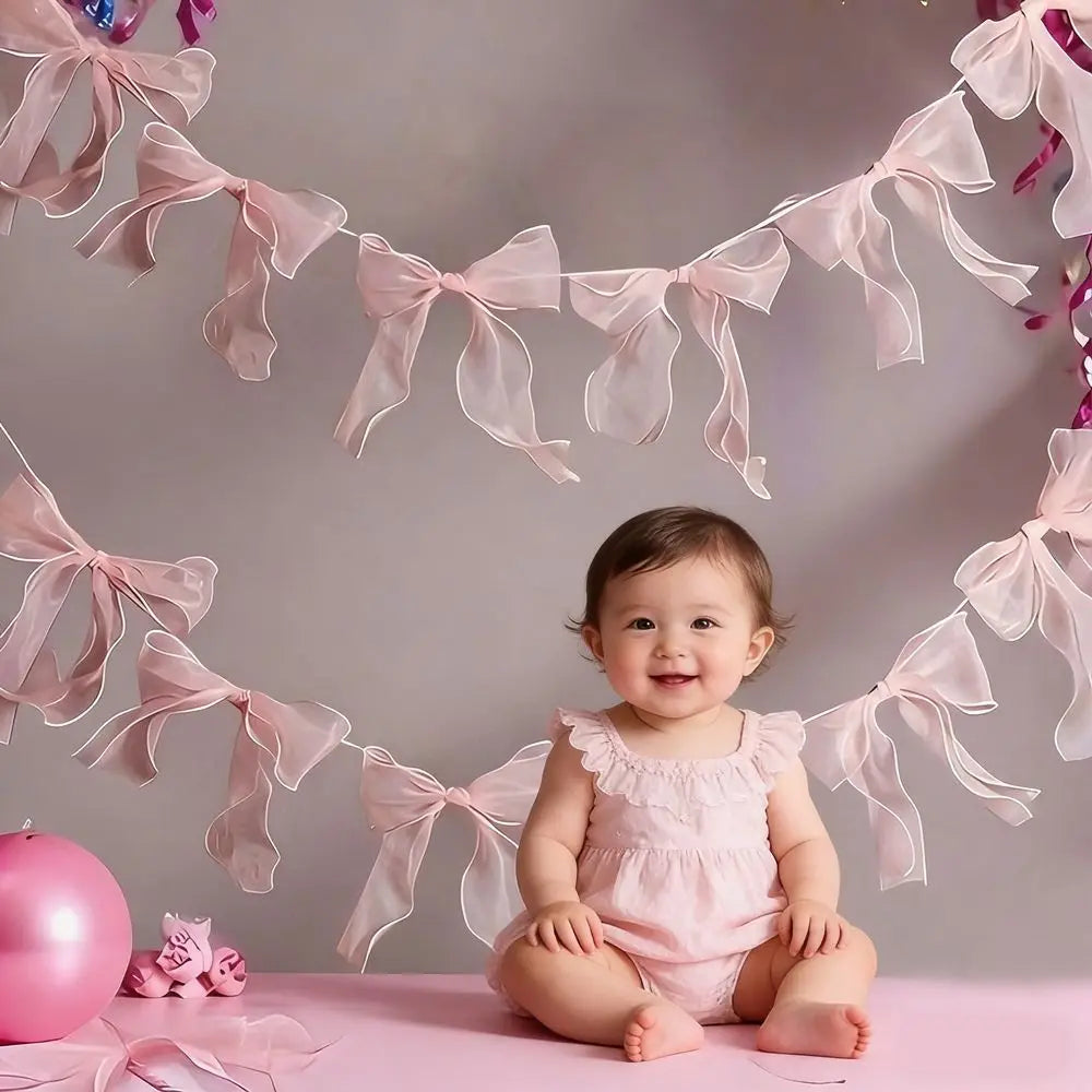 A baby girl sitting on a pink surface surrounded by a decorative bowknot banner garland set.