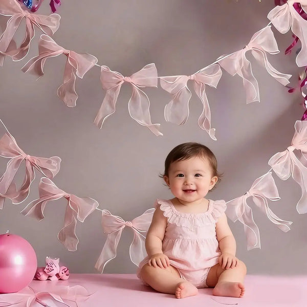 A baby girl sitting on a pink surface surrounded by a decorative bowknot banner garland set.