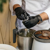 A person wearing black nitrile gloves is using a spoon to clean or handle a metal pot in a kitchen setting.