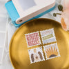 Goldener Teller mit Briefmarken, die Buchstaben und eine Sonnenform zeigen, neben einem weiß-blauen Stempelhammer.