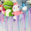 A colorful bohemian stuffed animal hammock featuring plush toys in pastel colors and purple, pink, and blue streamers.