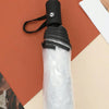 A transparent folding umbrella with a black handle and strap is shown against a terracotta background.
