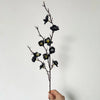 A slender branch of artificial plum blossoms with dark purple and yellow accents is held by a hand against a white background