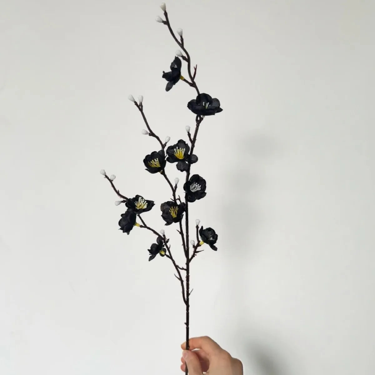 A slender branch of artificial plum blossoms with dark purple and yellow accents is held by a hand against a white background