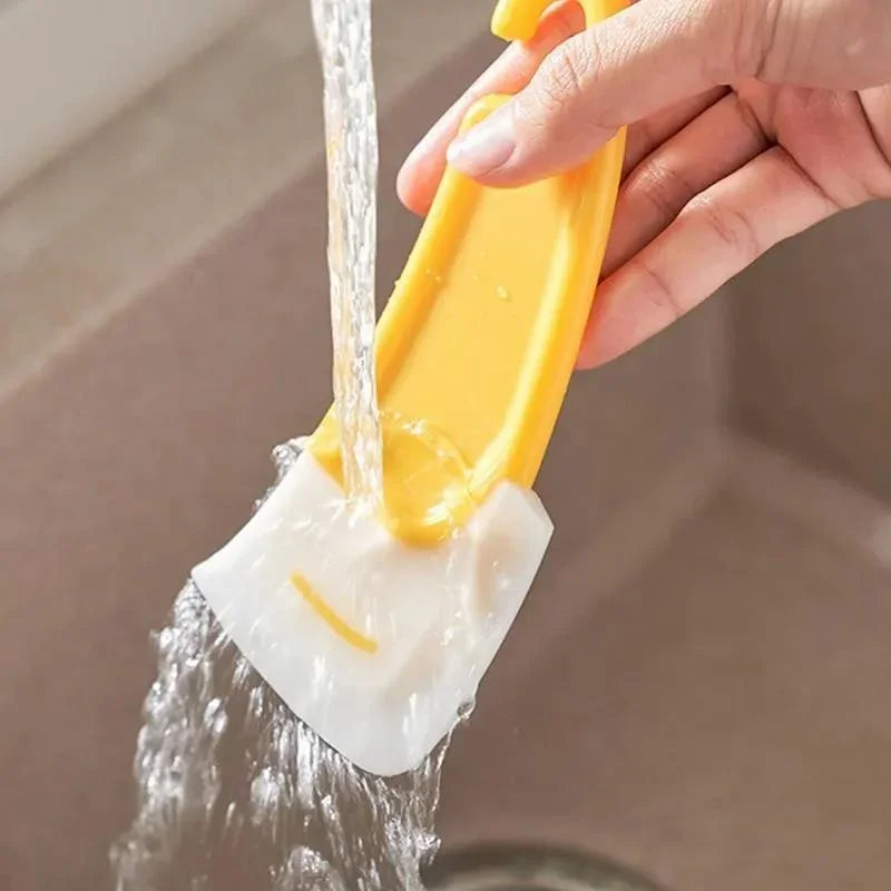 A hand is using a silicone kitchen scraper to wash a dish under running water.
