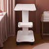A white portable beauty trolley cart with a flat top and storage shelf is shown in a softly lit room.