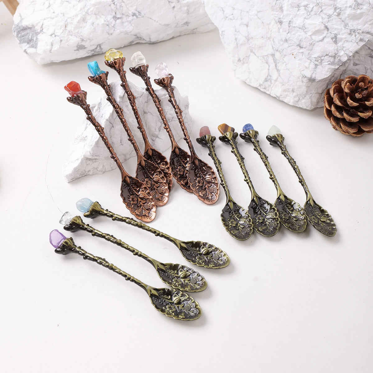 A collection of natural crystal spoons with various colors and shapes resting on a white surface next to a pine cone.