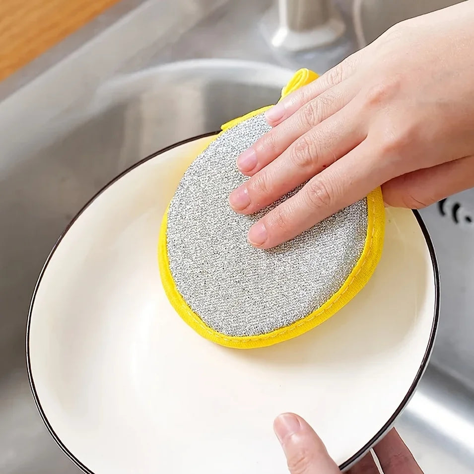 A hand is holding a yellow and silver reusable double-sided dishwashing sponge set over a white plate.
