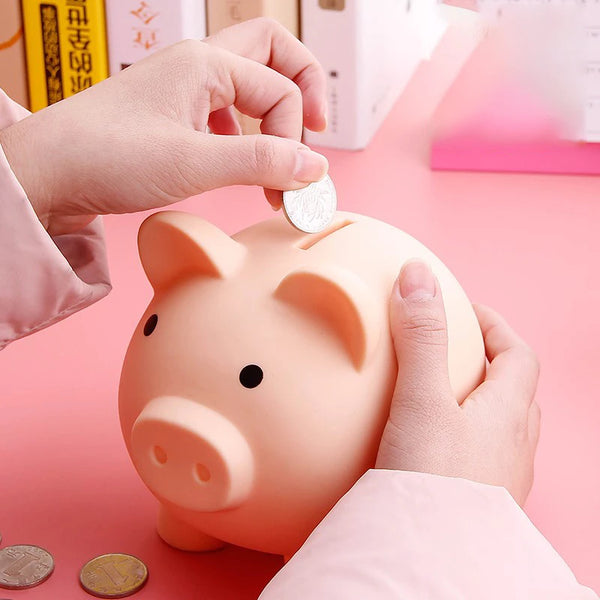 A hand is placing a coin into a pink vinyl piggy bank for kids on a pink surface.