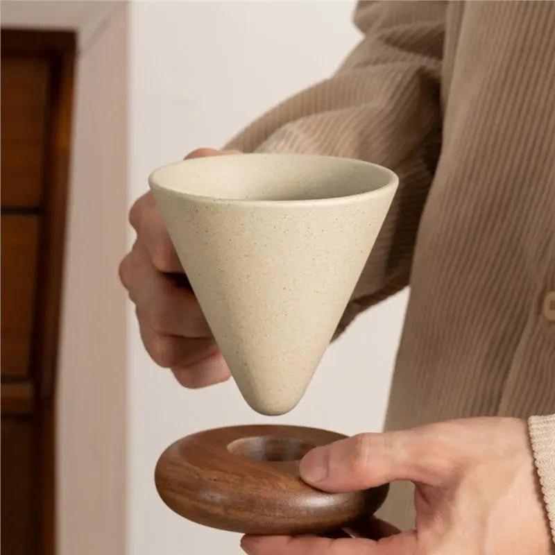 A person holds a light beige ceramic coffee mug with a wooden base and spoon set.