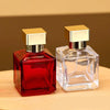 A clear glass perfume bottle with a gold cap and a red glass bottle are displayed on a wooden surface.